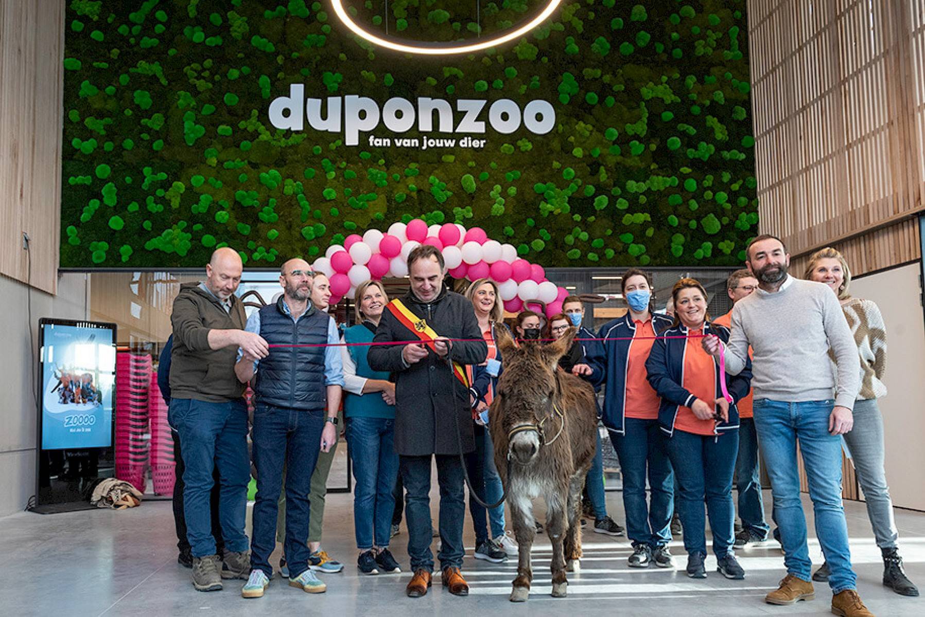 Medewerkers van Duponzoo poseren samen met een ezel, terwijl iemand een feestelijk lint doorknipt voor de ingang van Duponzoo